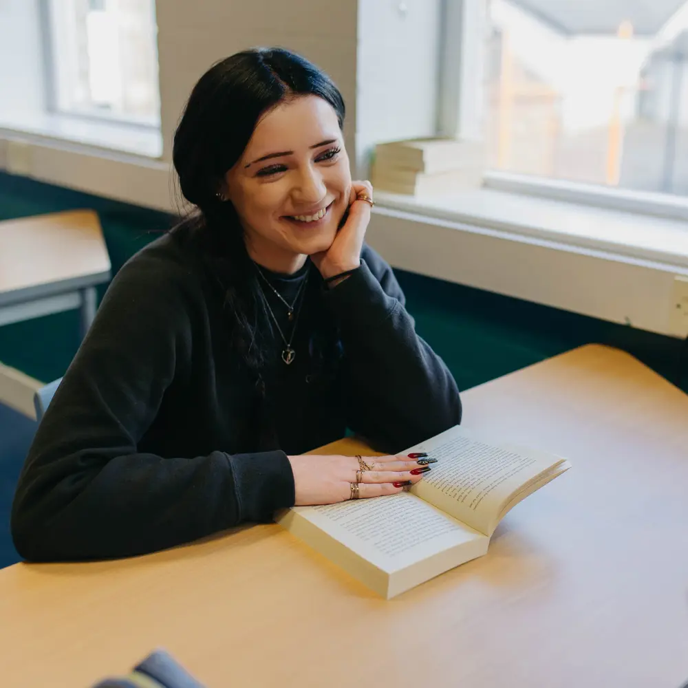 A young girl at Cedar House School with black hair smiling whilst reading a book