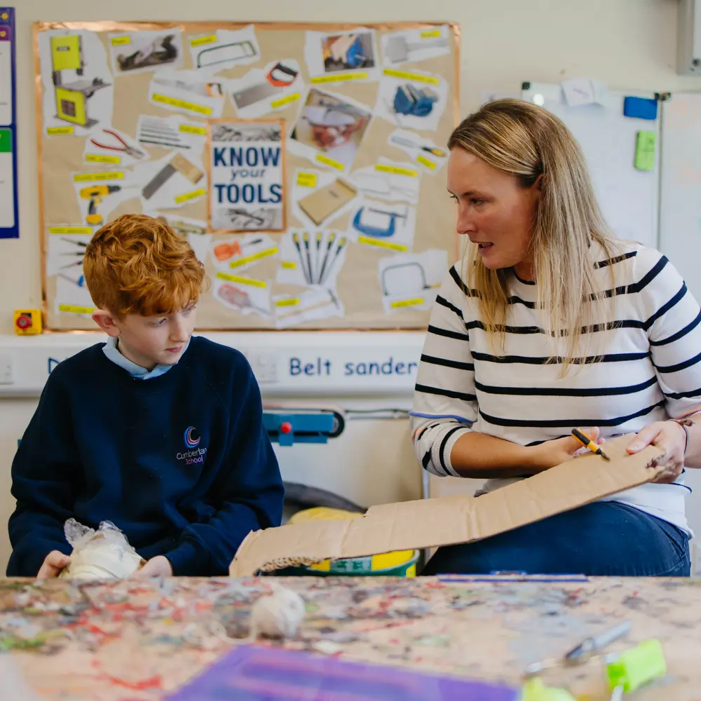Young boy and teacher from Cumberland School sat at a desk cutting cardboard