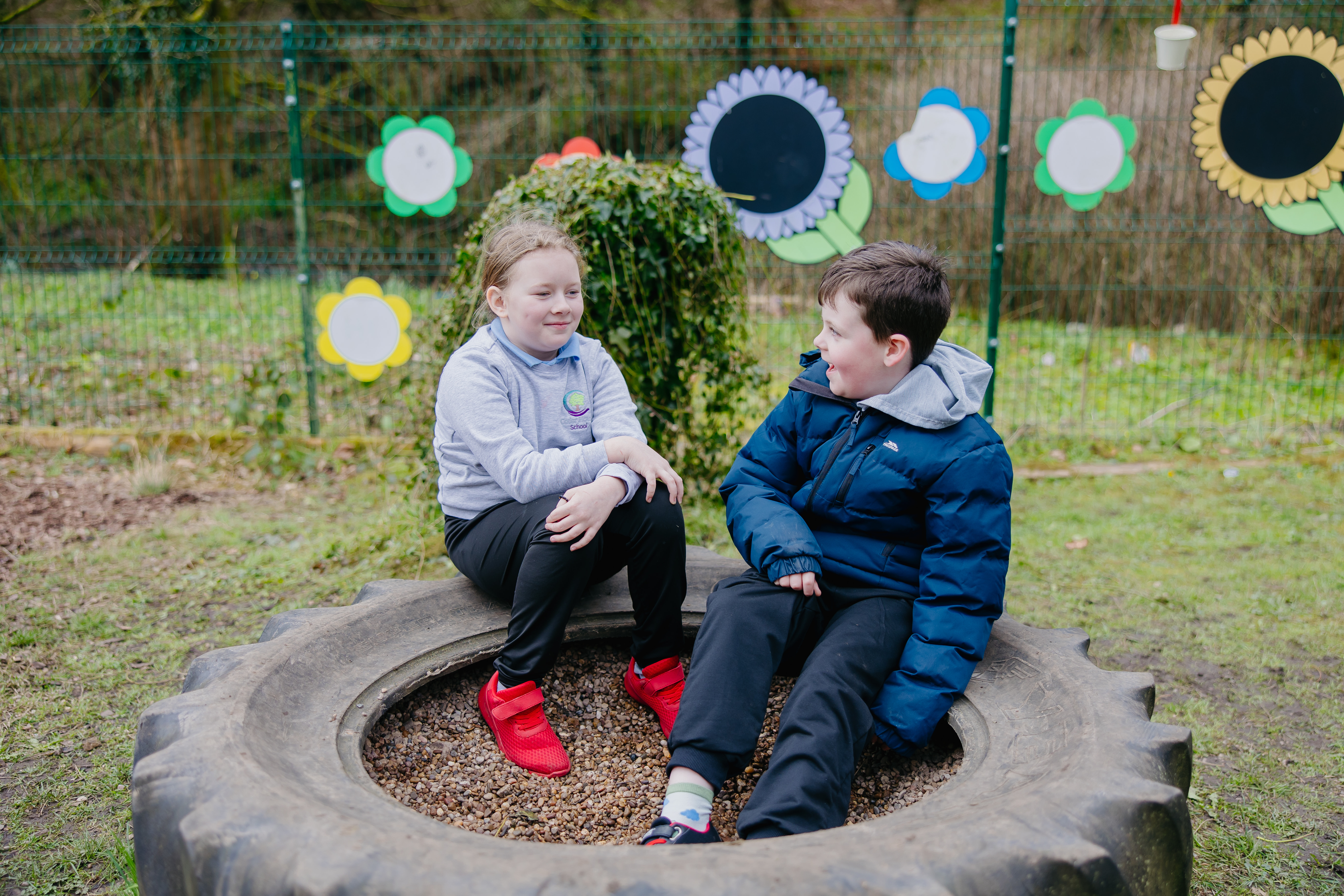 Two children, one boy, one girl, sat on ede of a tyre plant bed 