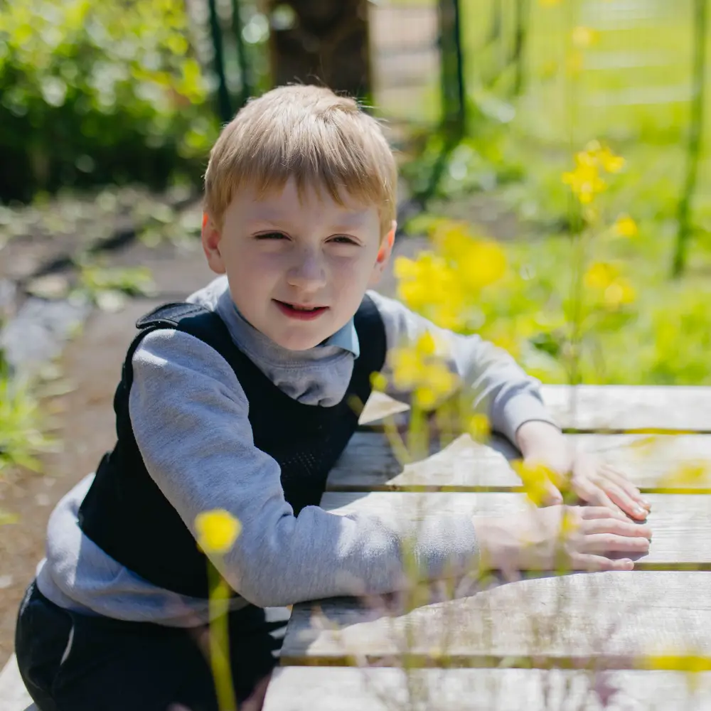 Young boy from Cedar House School sat on a bench outside surrounded by yellow flowers smiling at camera