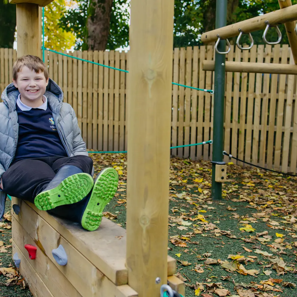 Boy in blue school uniform and grey coat sat on a wooden playground