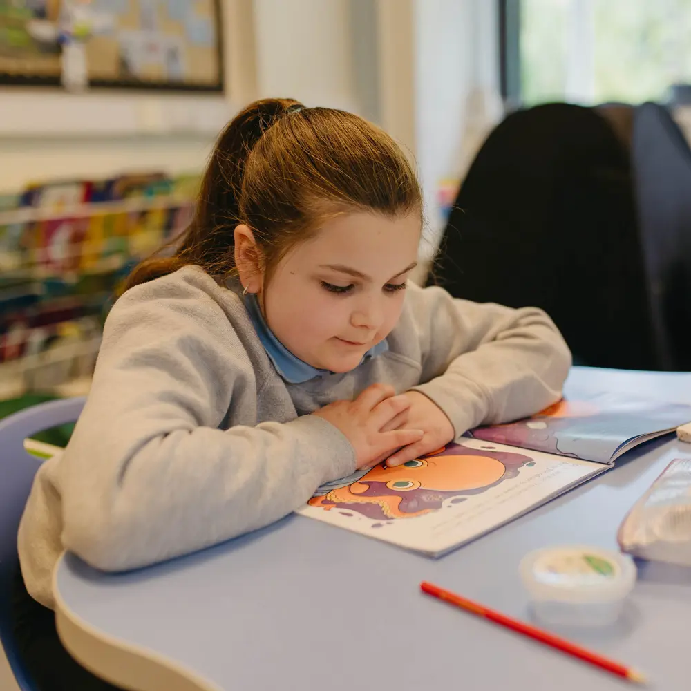 Young girl from Cedar House School reading a book at a desk in a classroom