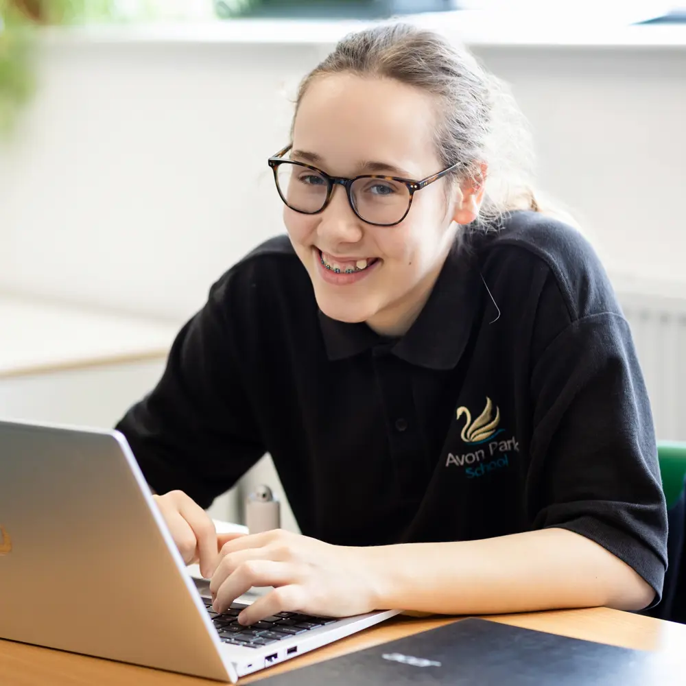 A young girl with glasses at Avon Park school smiling at the camera whilst on a laptop