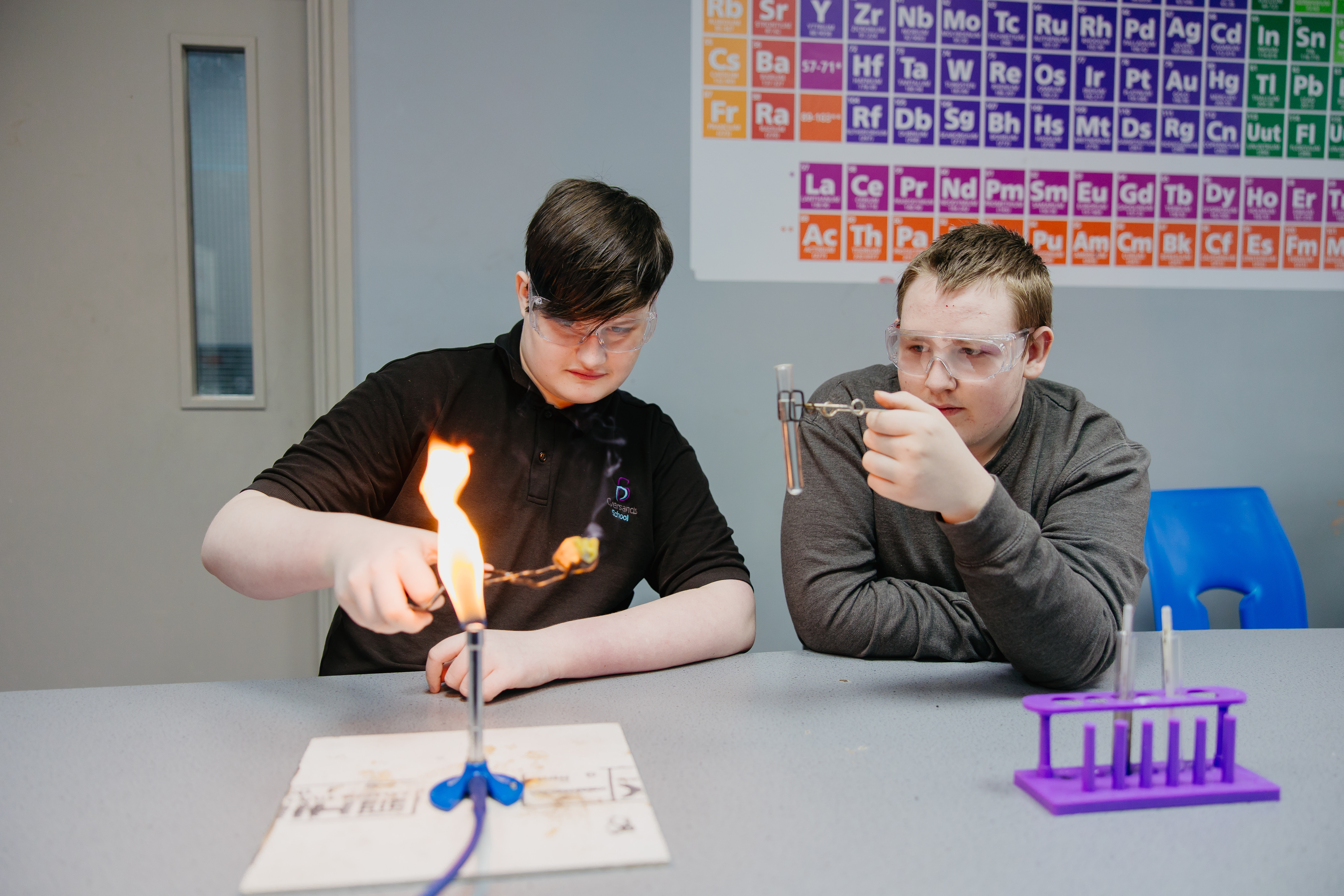 Two boys sat next to eachother wearing goggles perfoming science experiment with test tube and bunsen burner
