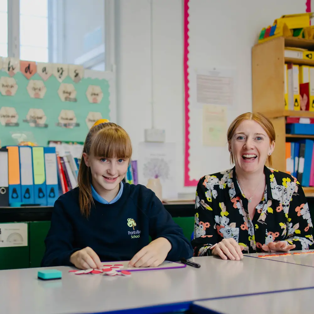 Girl and teacher sat at a desk in a classroom at Pontville School smiling into the camera