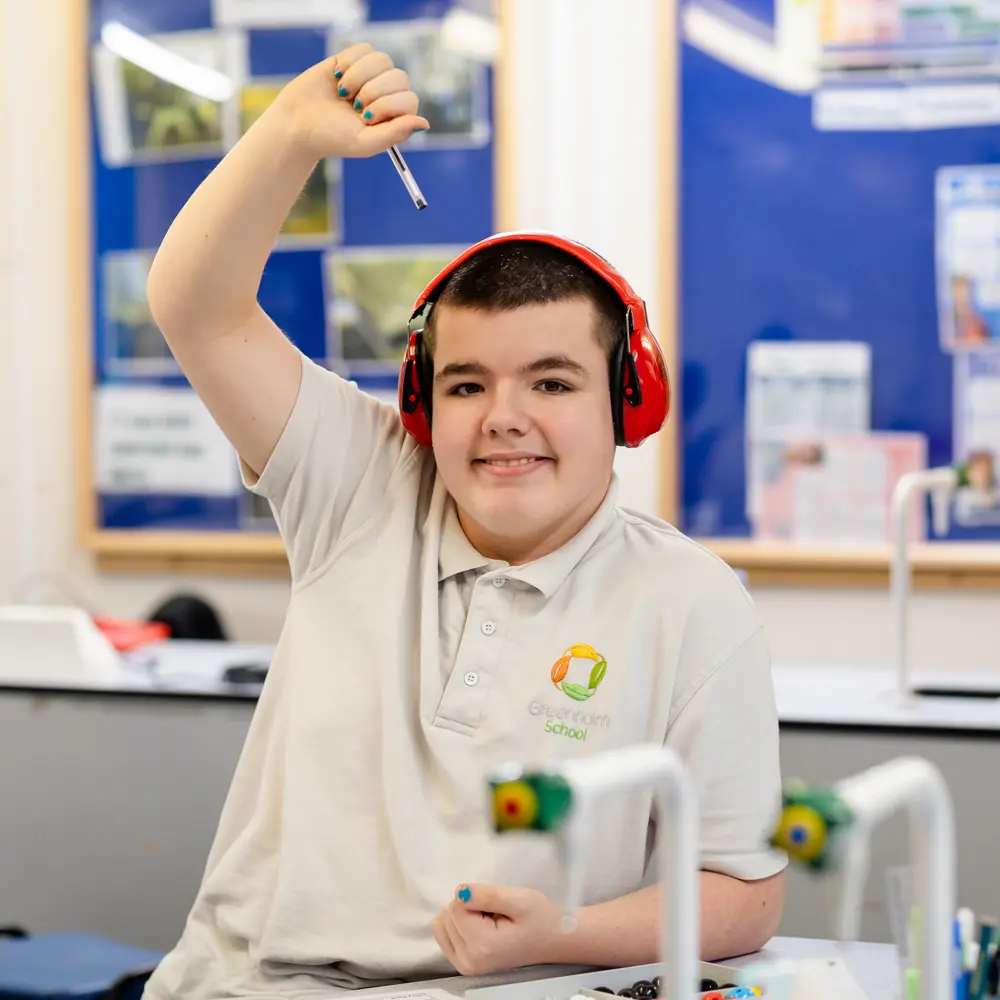 Boy from Greenholm School sat in class with ear defenders on with his hand raised