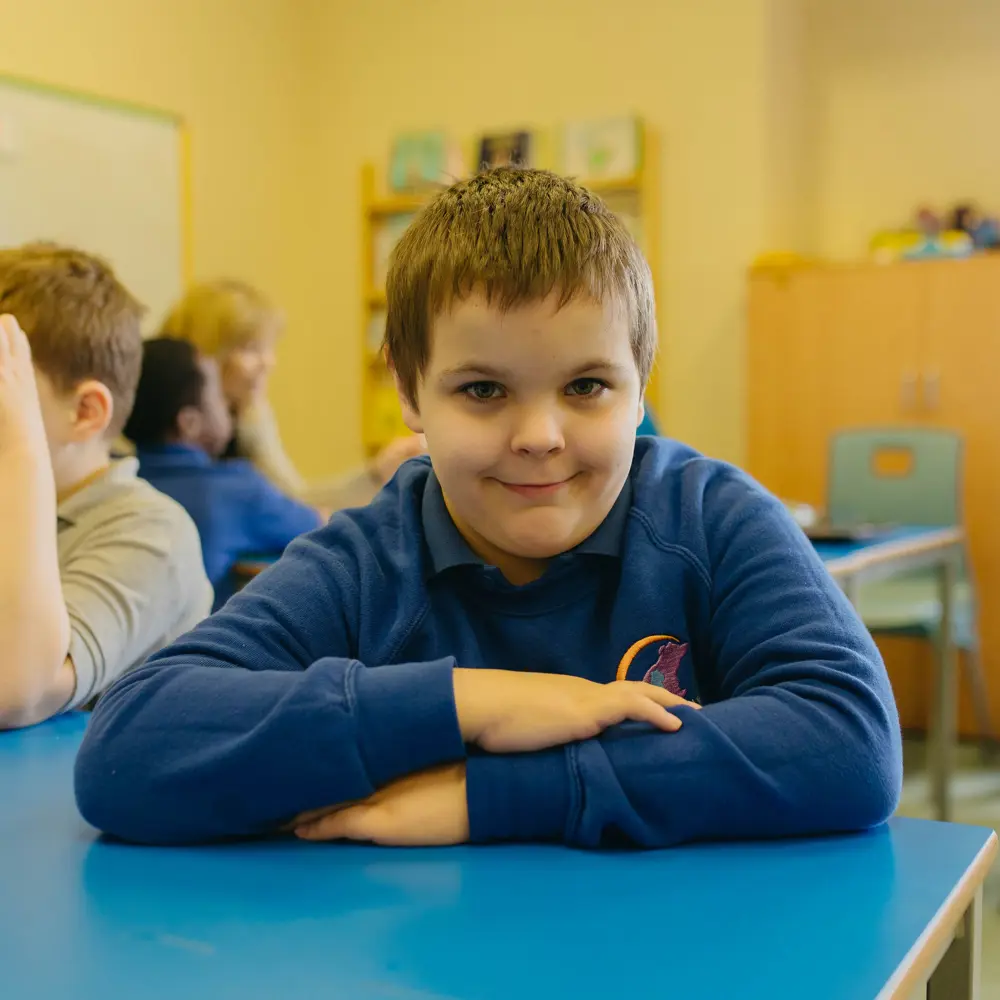 A young boy at Westmorland School sitting in a classroom with his arms crossed on the table smiling at the camera