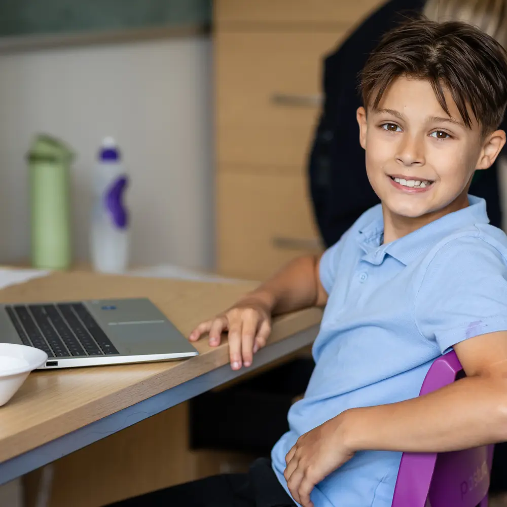 Young boy with brown hair from Hinckley House School smiling into the camera while working on his laptop
