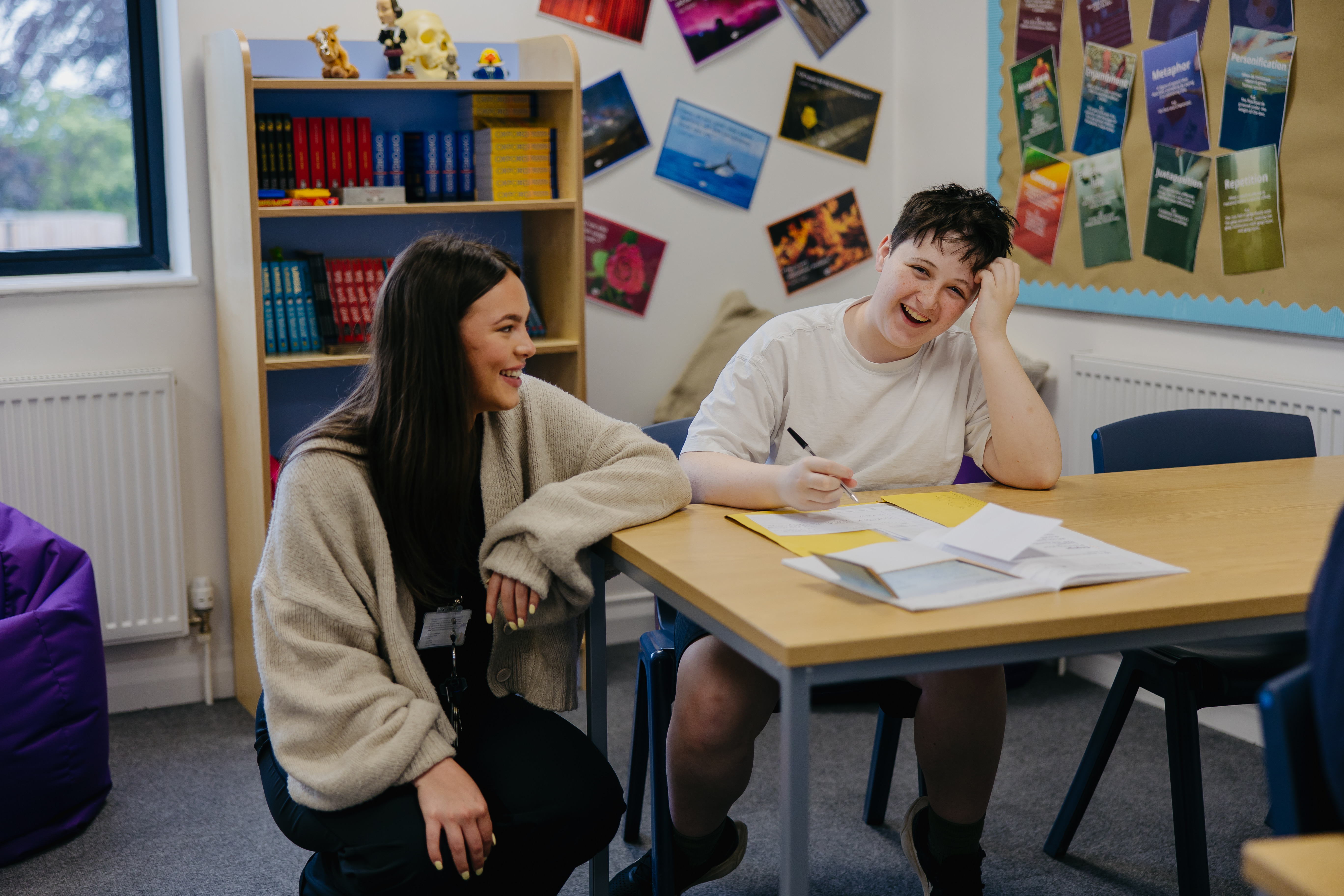 Pupil sat at desk laughing and smiling as teacher crouches next to them