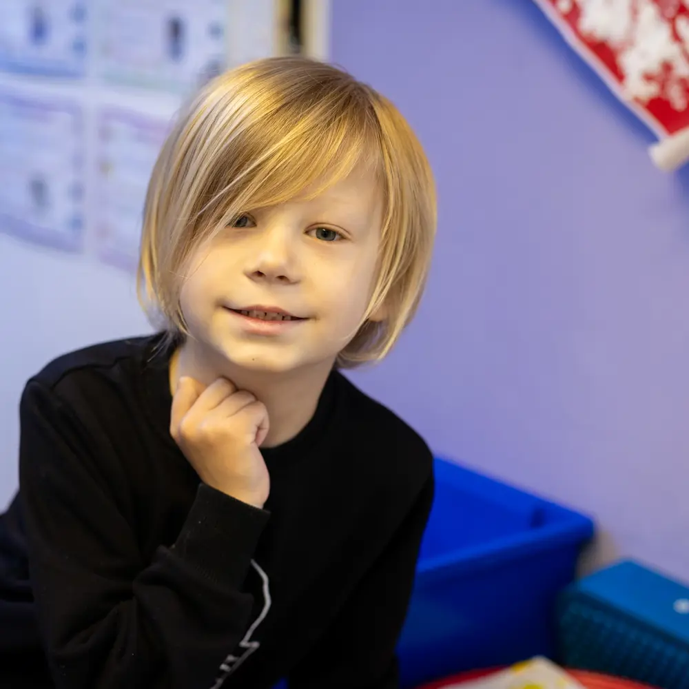 A young boy with blonde hair at Lavender Lodge School smiling at the camera in a classroom