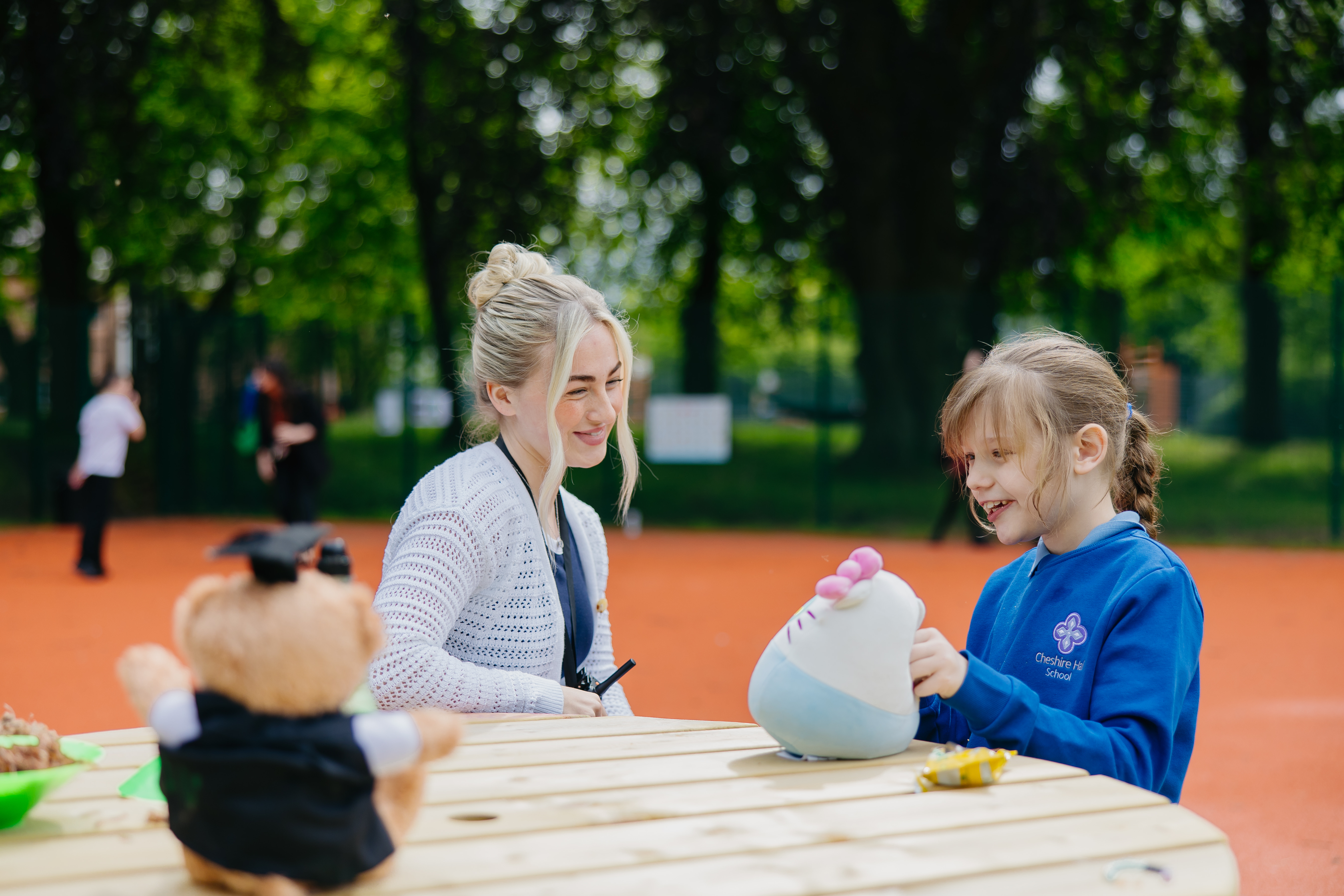Girl and staff member sat at bench outside in play area playing with soft toys