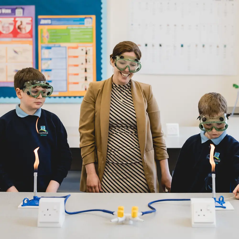 Two pupils and a teacher in a science lesson using Bunsen burners wearing protective glasses