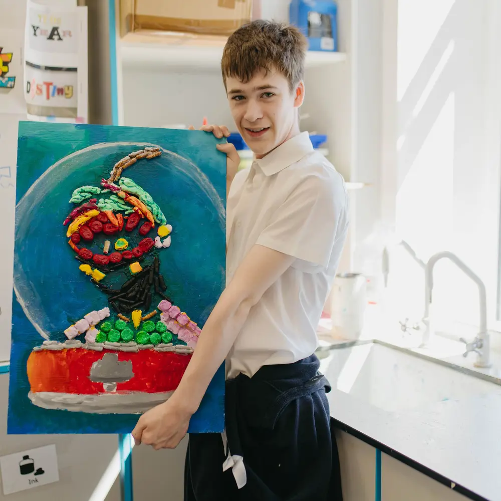 Young boy from Hartwell School with brown hair shows off artwork in classroom