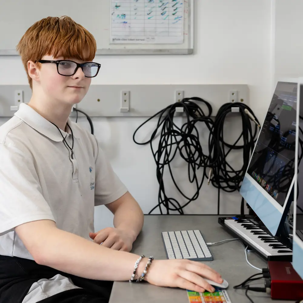 A young boy with red hair at Dovetree School sitting at a computer desk looking at the camera