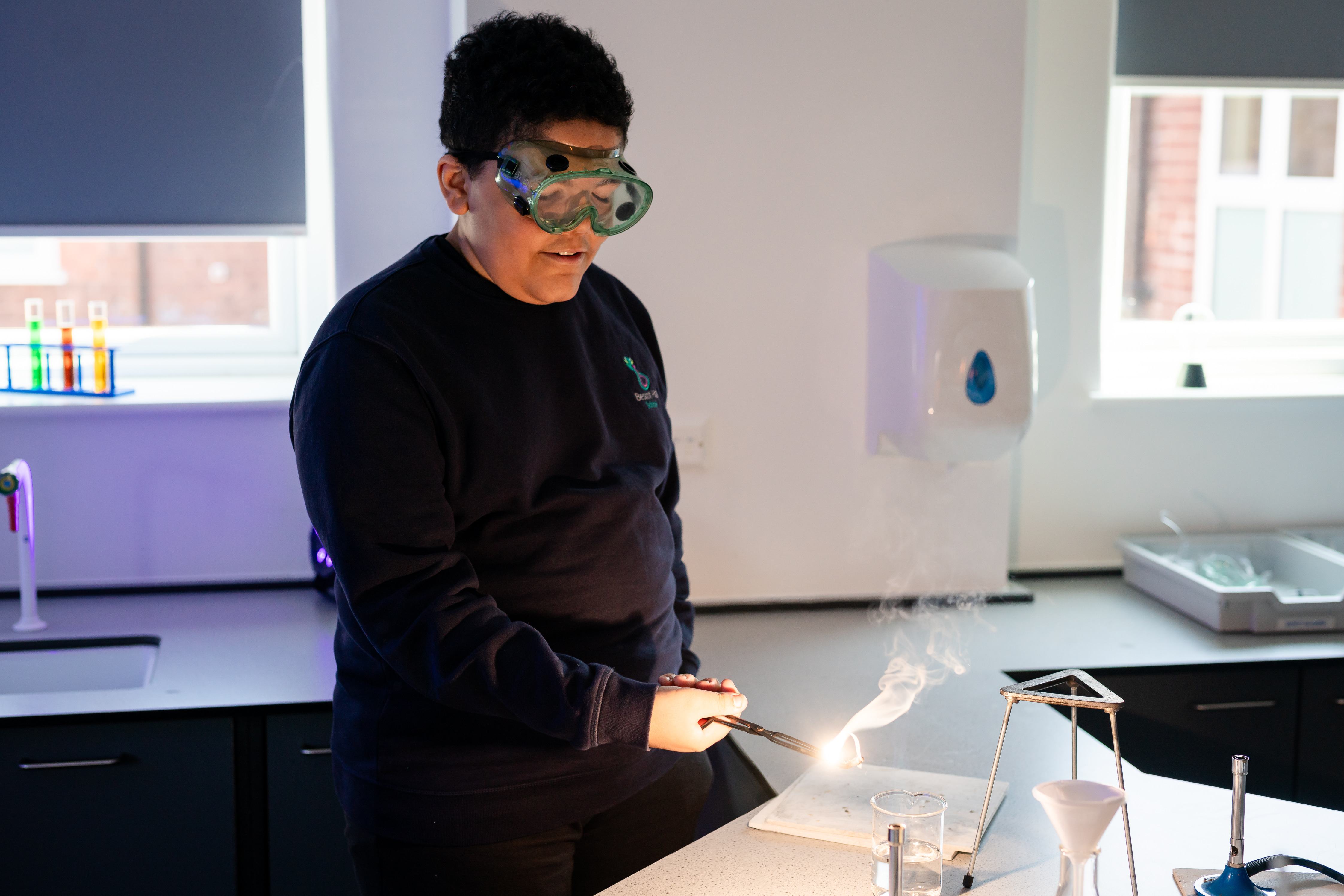 Boy wearing black jumper and science goggles using bunsen burner