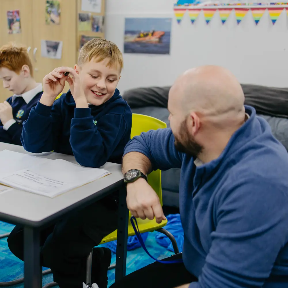 2 boys sat at a desk in a classroom engaging with a male teacher