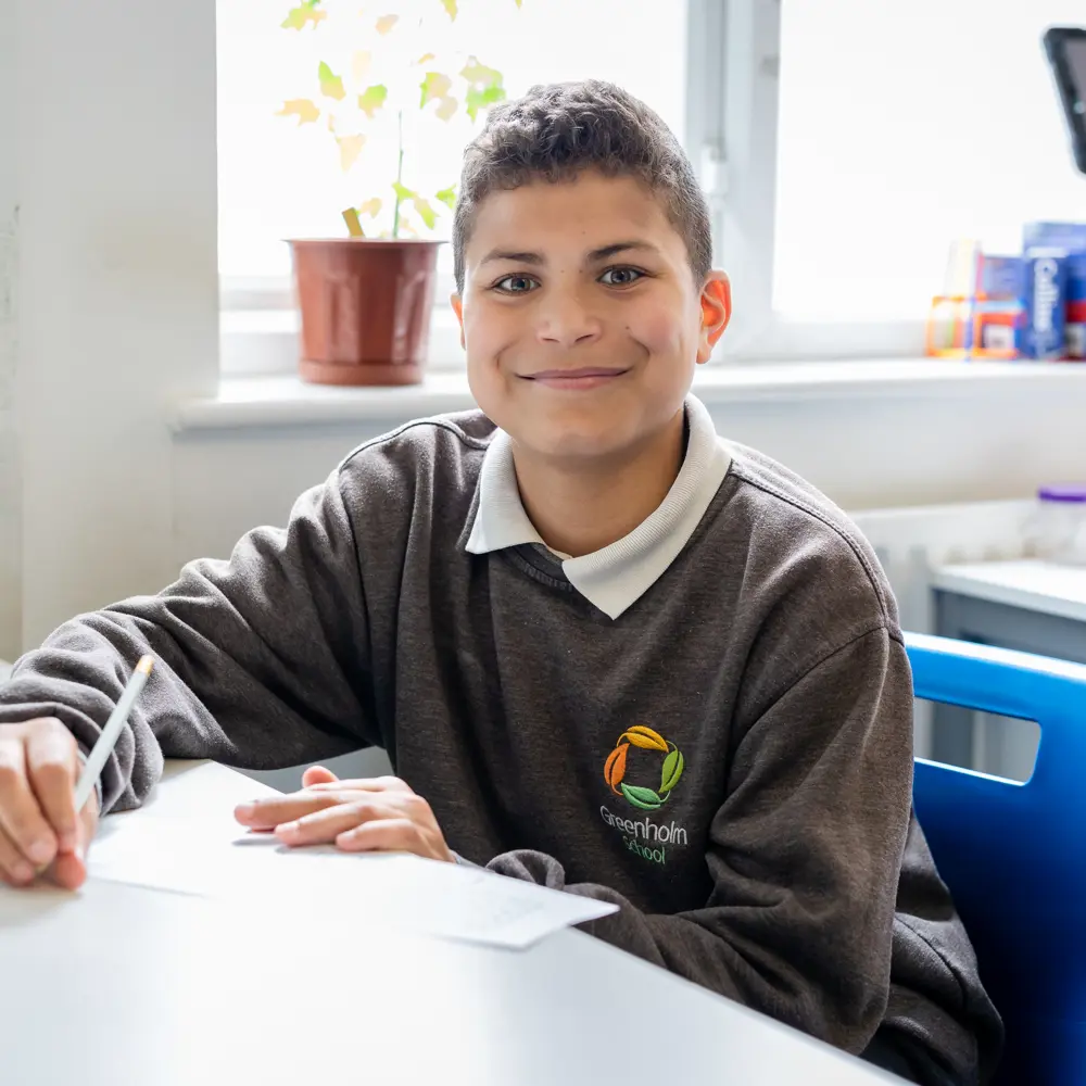 A young boy at Greenholm School sat at a desk working smiling at the camera