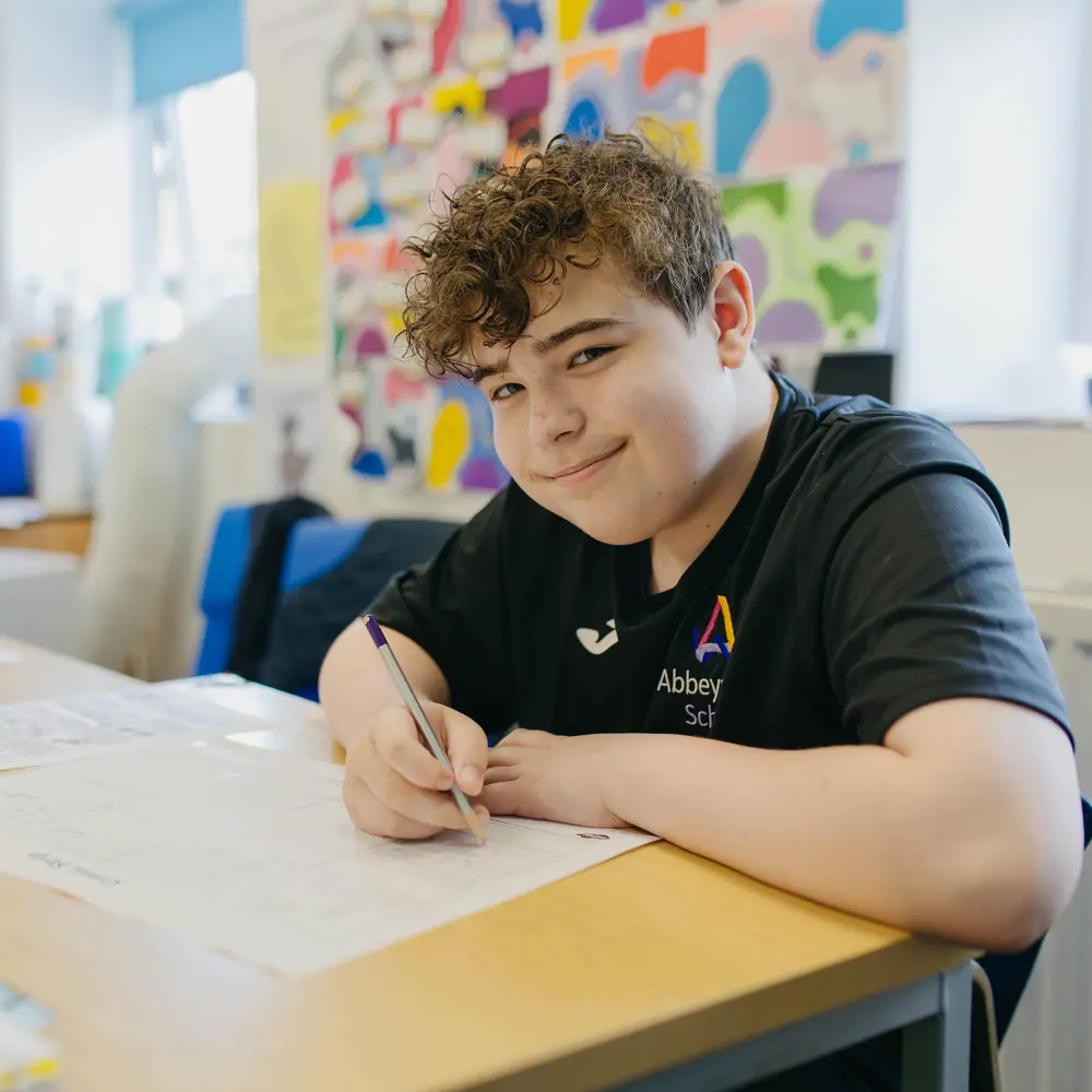 Smiling brown-haired boy sat a desk completing classwork in a classroom at Abbeywood School