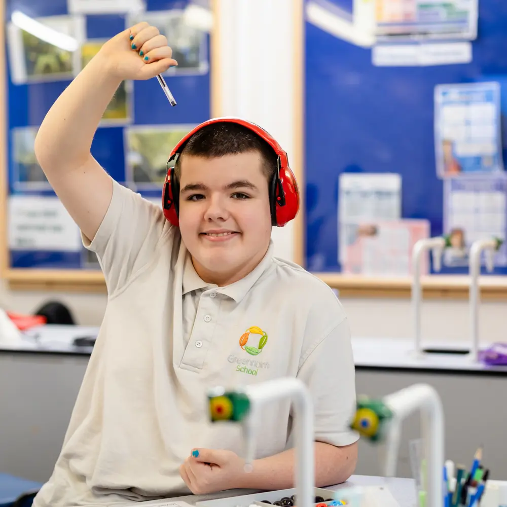 A young boy at Greenholm School in a science lesson wearing ear defenders with his hand raised