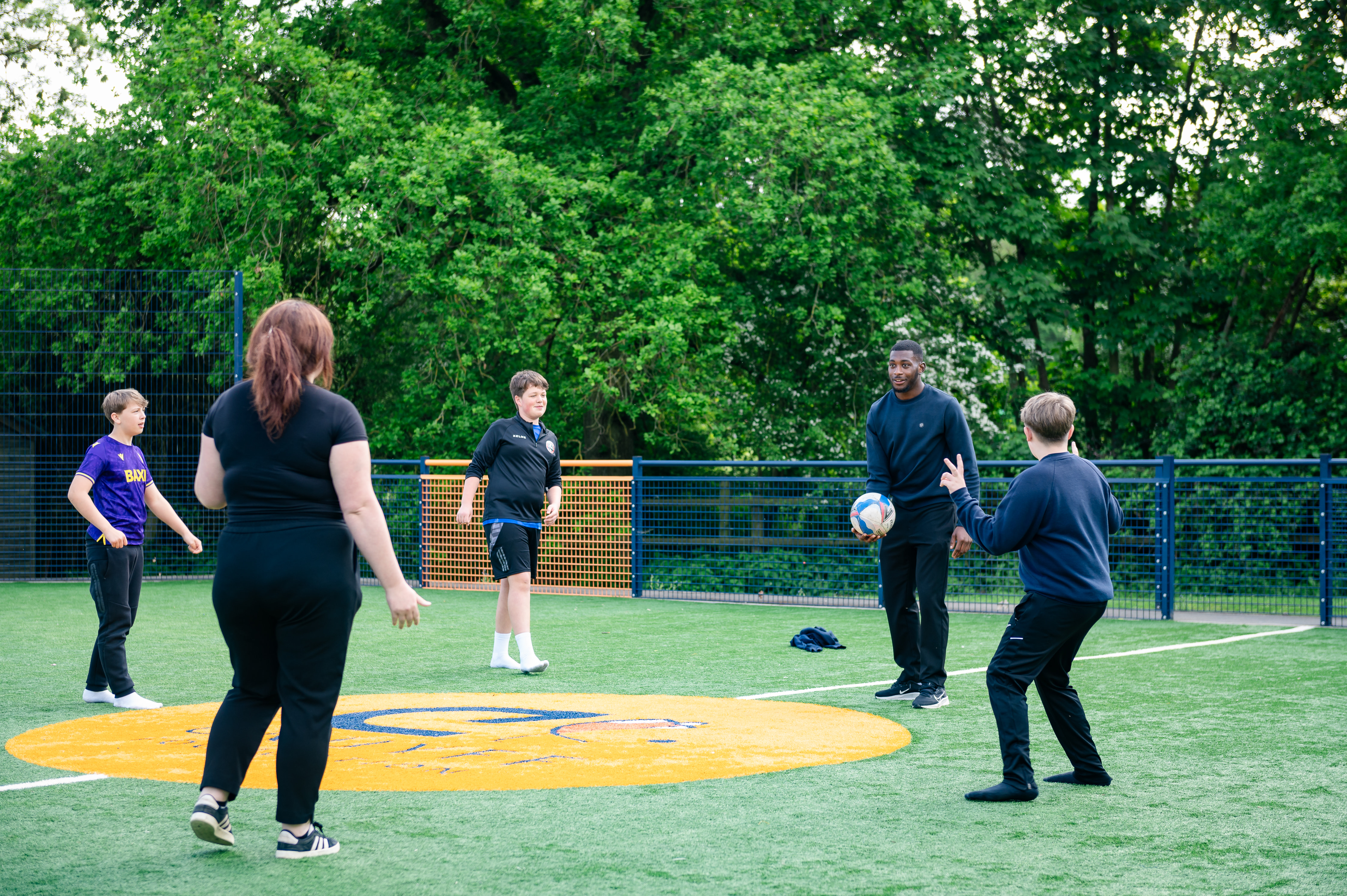 Three pupils and two members of staff stood in circle passing round a football outside on astroturf