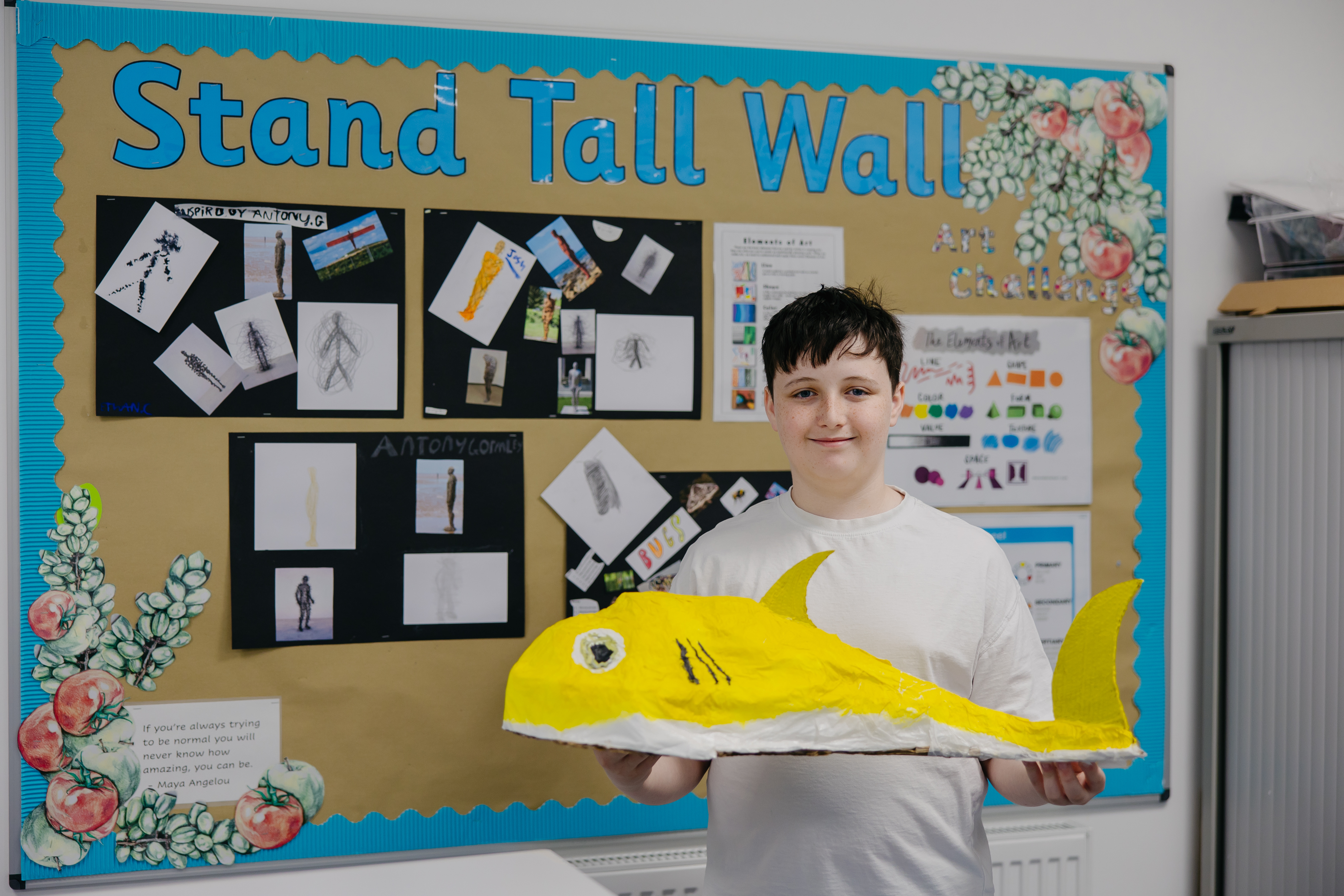 Boy stood infront of display board while holding big handmade art piece of fish