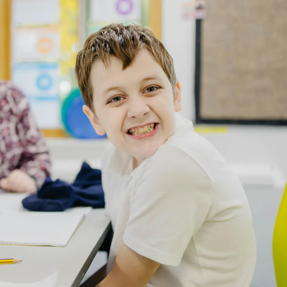 Smiling brown-haired boy sat at a desk doing his classwork with the help from a female teacher