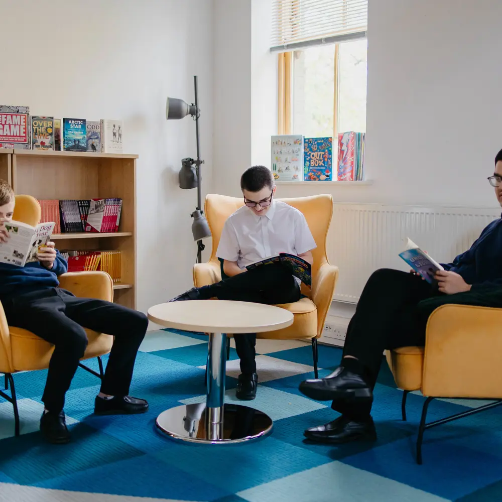 3 boys sat on yellow chairs reading books in the school library