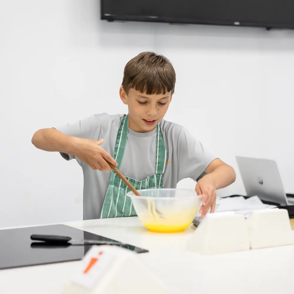 A young boy in a grey t-shirt and a green apron mixing cake ingredients with a wooden spoon at Queensmead House School
