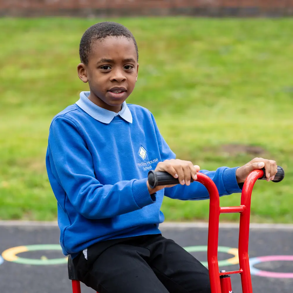 Young boy from Hinckley House School sat on a red bike in the playground looking at the camera