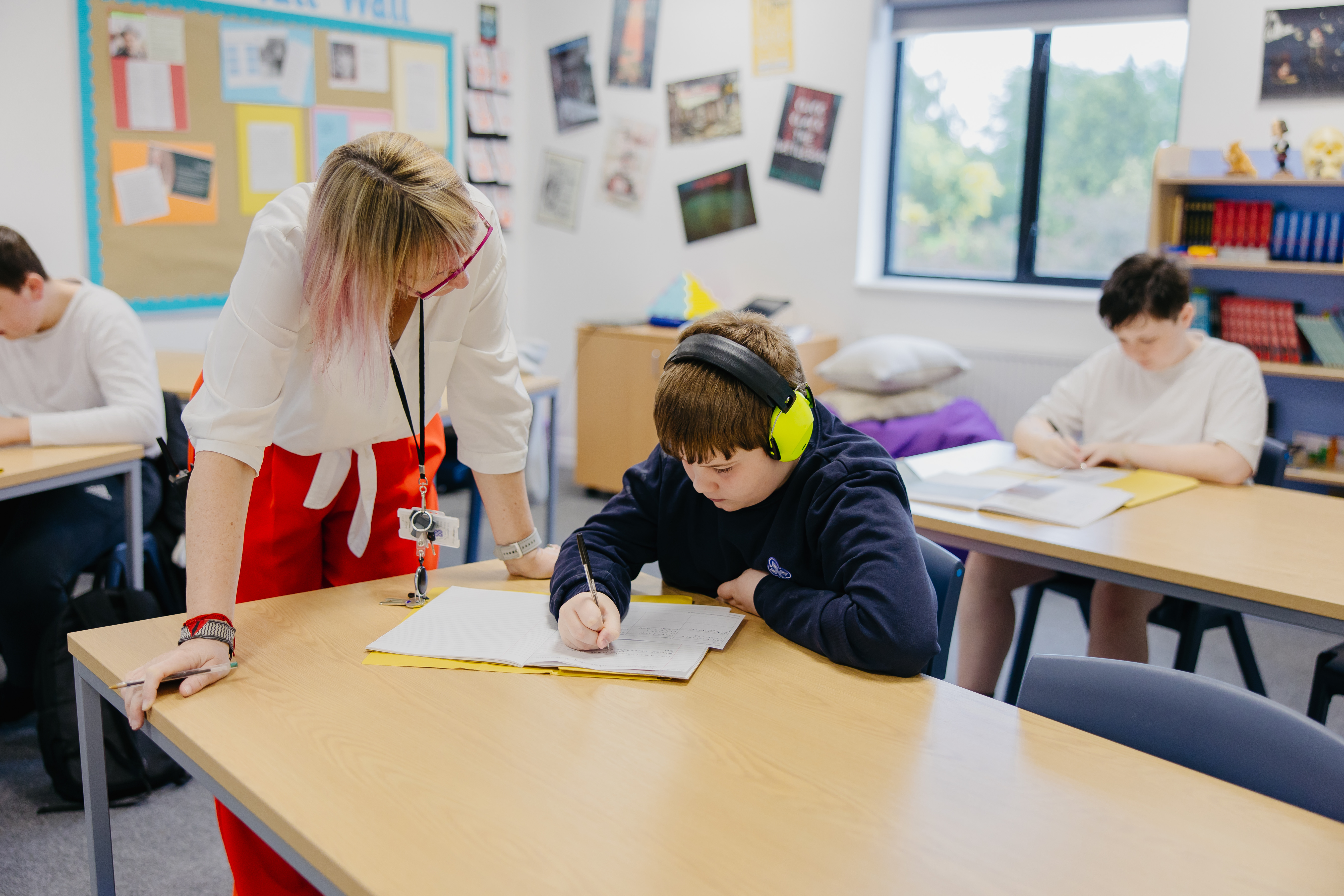 Boy sat at desk doing school work while teacher stand newt to him