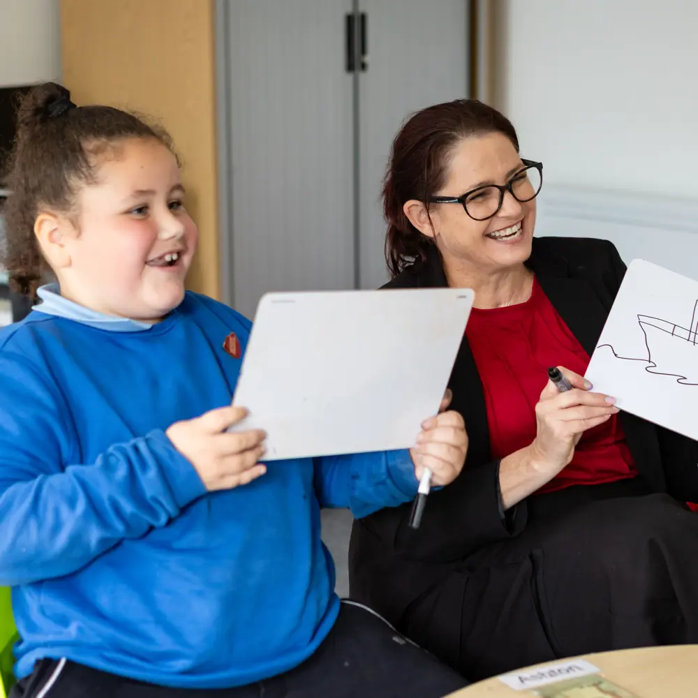 Young pupil with teacher at Hinckley House School in classroom holding up whiteboards with drawings on