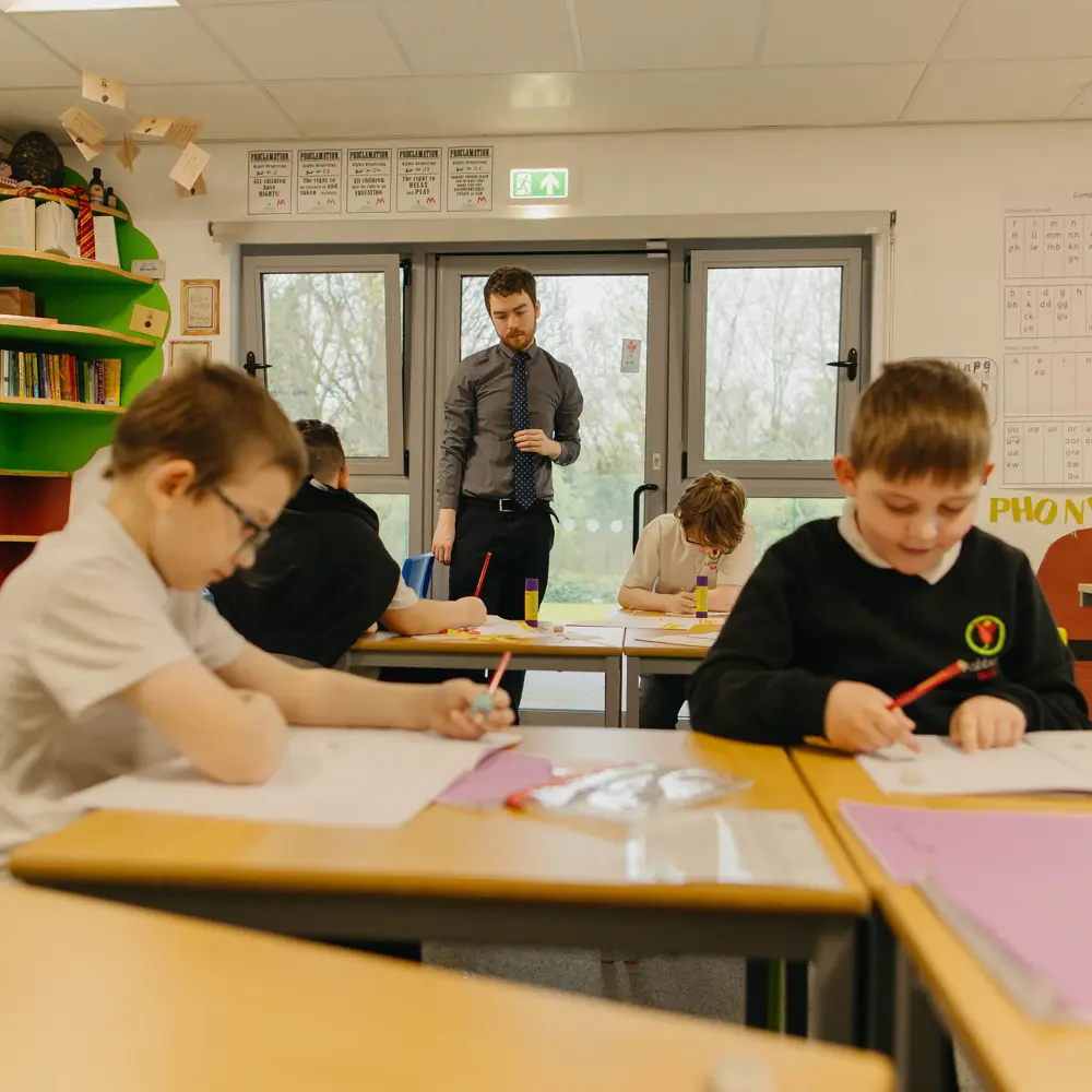 4 boys sat at a their desks holding pencils and completing classwork with a male teacher observing