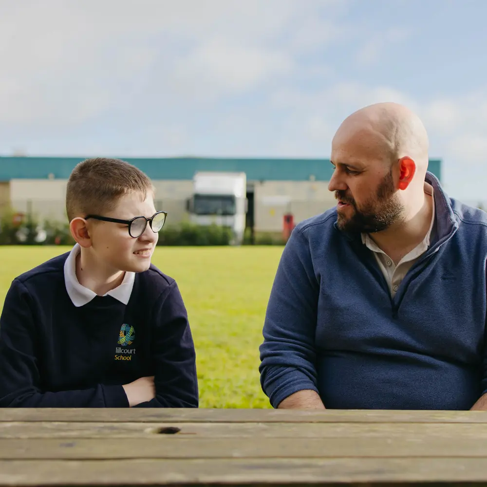 A young boy with glasses at Millcourt School sat on a bench with a member of staff
