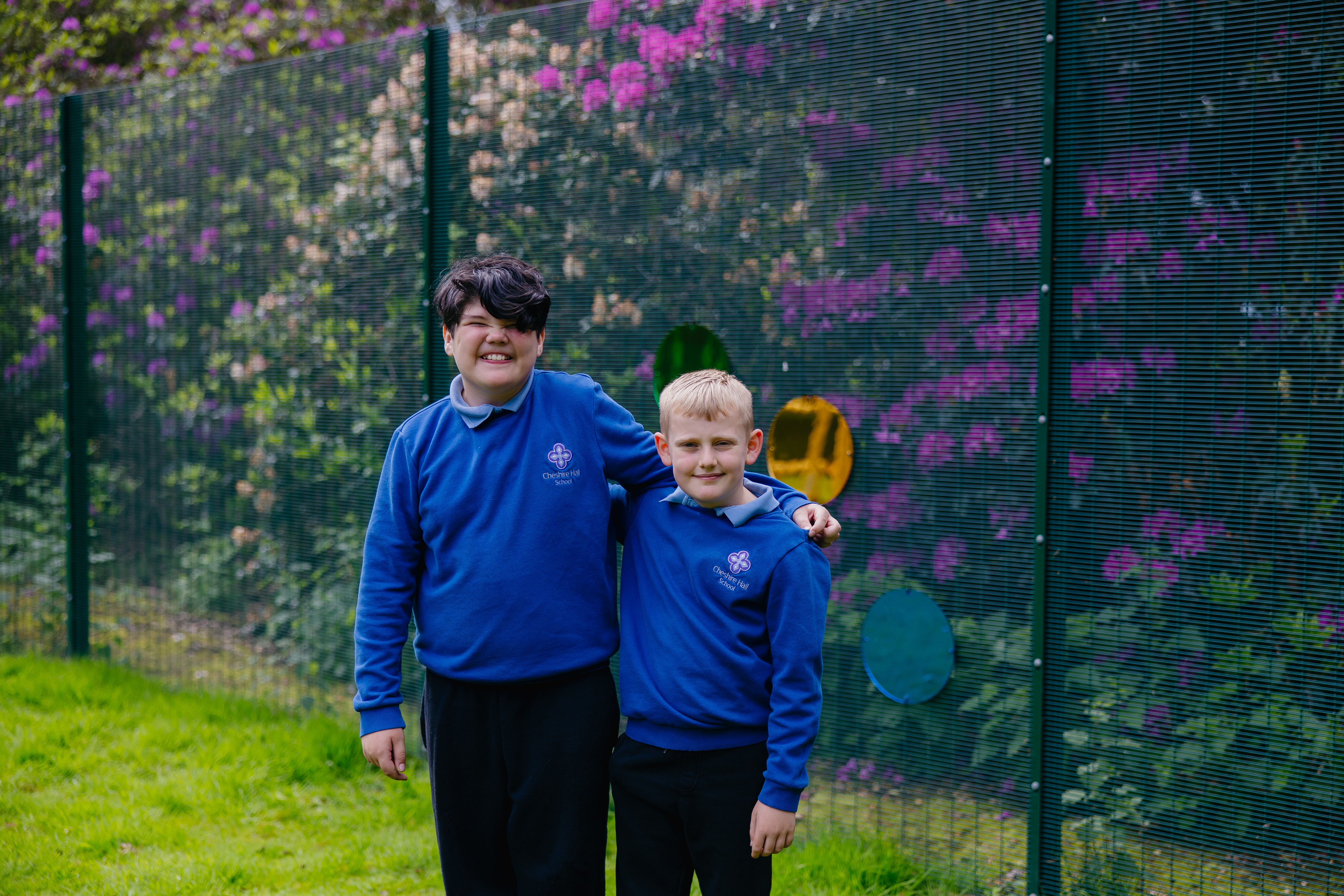 Two boys stood with arms around eachother smiling and lookin at camera